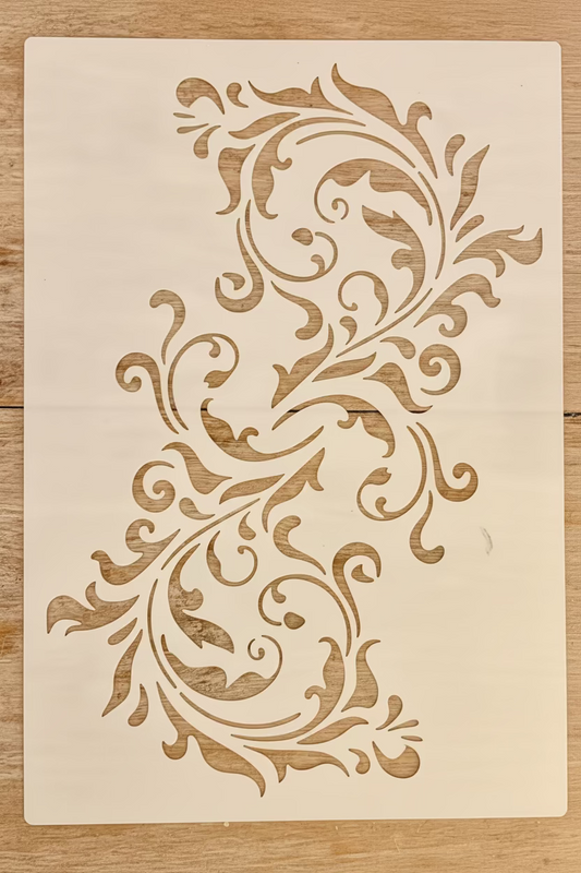 A rectangular white stencil or template sheet featuring a symmetrical, vertical baroque-style scroll and leaf pattern, displayed flat on a light brown wooden surface.